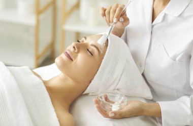 Kozmetik uzmanı, spa salonundaki genç, rahat, gülümseyen bir kadına nemlendirici maske takıyor. Güzellik kliniğindeki güzellik uzmanından güzellik prosedürü alan bir kız müşterinin portresini yakından çek