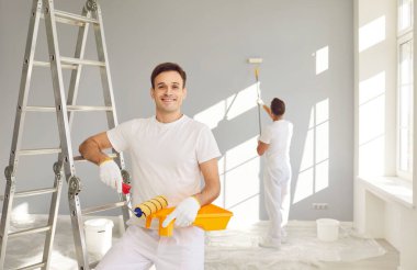 Erkek işçiler apartmanda iç yenileme yapıyorlar. Mutlu gülümseyen, kendine güvenen, beyaz üniformalı, elinde boya silindiri ve tepsisiyle kameraya bakan yetişkin duvar ressamı portresi.