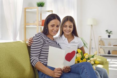 Genç anne ve kız evdeki kanepede oturup birlikte tebrik kartı okuyorlar. Anneler ve Kadınlar Günü için bir buket çiçek lalesiyle annemi kutlayan mutlu kız çocuğu..
