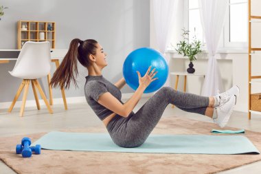 Evdeki oturma odasında, formda bir topla yoga minderinin üzerinde uzanan sıska bir kadın. Spor kızı içeride egzersiz yapıyor. Sağlıklı yaşam tarzı, spor ve spor anlayışı.