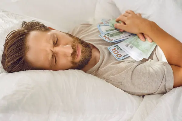 Worried man sleeping with his money savings on chest. Adult bearded man in bed on white pillow with bunch of dollar bills in hands dreaming bad dreams with facial expression of fear. Crisis concept