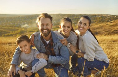Çocuklu aile birbirine sıkıca sarılıp altın saatinde kaygısız anın tadını çıkarıyor. Baba, anne, oğul ve kızın portresi güzel doğal manzaranın arka planında gülümsüyor..