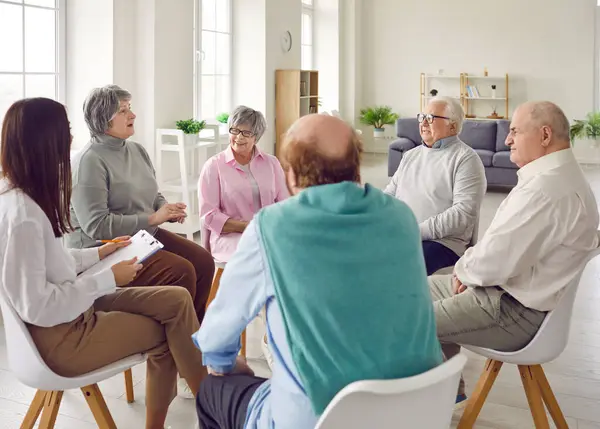 Przyjazna Młoda Pielęgniarka Opiekunka Lub Psycholog Rozmawiająca Grupą Seniorów Mężczyzn Zdjęcie Stockowe