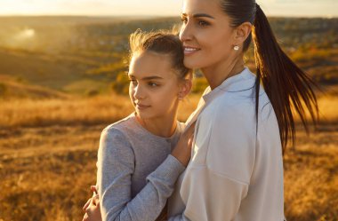 Genç gülümseyen anne dışarıda mutlu kızıyla birlikte tarlada duruyor. Doğanın tadını çıkaran bir kadın ve genç bir kız mesafeye bakarlar. Aşk, ilgi ve anneler günü konsepti.