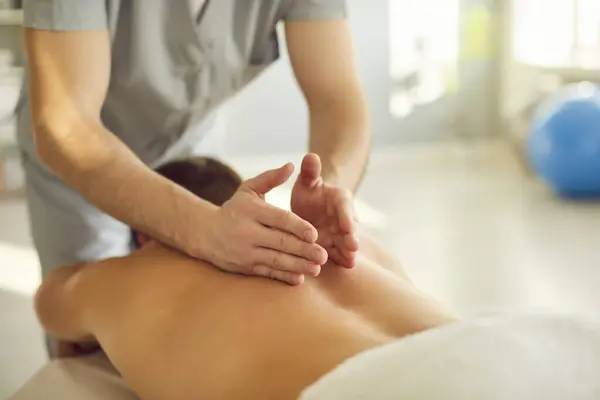 Unrecognizable professional masseur doing remedial, holistic, relaxing, healing body massage for client. Close up specialists hands on young mans back. Massage, therapy, health concept background