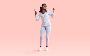 Genç, neşeli, Afro-Amerikan bir kadının portresi. Günlük giysiler içinde dans ediyor ve kulaklıkla müzik dinliyor. Cep telefonu stüdyoda pembe bir arka planda..