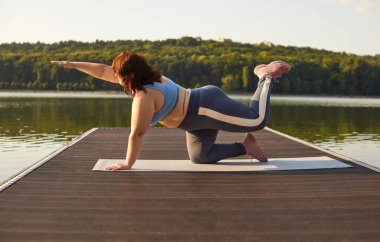 Spor giyimli şişman bir kadın rıhtımdaki yaz parkında spor egzersizleri yapıyor. Kilolu bir kız yoga minderinde dışarıda egzersiz yapıyor. Vücut pozitifliği ve açık hava eğitim kavramı