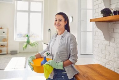 Genç, mutlu, gülümseyen bir ev kadınının portresi lastik eldiven takmış, temizlik aletleri ve paçavralarla modern mutfağın içinde duruyor ve kameraya neşeli bakıyor. Ev işleri ya da ev işleri..