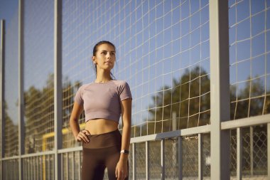 Çekici bir kadın model spor çitlerinin arkasında el ele poz veriyor. İnce vücutlu güzel bir kadın tişört giyiyor açık havada mavi gökyüzünün altında sabah sporu yapıyor.