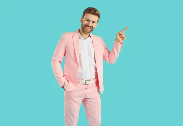 快乐的男人指着身边 穿着粉红西服的英俊年轻人手插在蓝色背景的口袋里 展示着什么 登广告推销时尚 微笑着把手指指向一边 图库照片