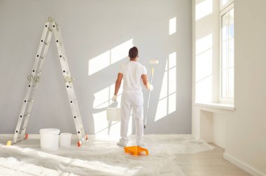 Beyaz elbiseli bir erkek ressam boş bir odada elinde bir kova ve gri bir duvarı boyamaya gidiyor. Yenileme, apartman onarımı ve yenileme konsepti..