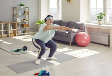 Mutlu şişman kadın evde aktif fitness egzersizi yapıyor. Spor kıyafetli neşeli şişman genç bayan oturma odasında bir spor paspasın üzerinde duruyor, çömeliyor ve gülümsüyor.