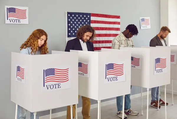 一群年轻而快乐的 多样化的美国公民的画像人们站在投票中心 举着美国国旗在投票亭里 在选举日把选票放进垃圾箱 图库图片