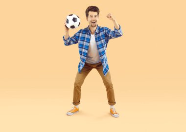 Neşeli heyecanlı adam dünya kupası maçında en sevdiği futbol takımını destekliyor. Futbol topu tutan ve bej arka planda yumruğunu sıkan komik yüz ifadesi olan adam. Futbol taraftarı kavramı.