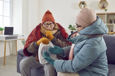 Son sınıf aile çifti evde çok üşüyor. Küçük elektrikli vantilatör ısıtıcısı ile ısınan kışlık montlu iki emekli yaşlı insan, soğuk, ısıtılmamış evde, oturma odasındaki kanepede birlikte oturuyorlar.