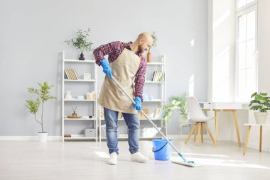 Önlük ve eldiven giymiş mutlu bir adam evi temizliyor ve yüzünde bir gülümsemeyle yerleri paspaslıyor. Neşeli tavırları ev işlerini tamamlamak ve temizlik yapmaktan keyif aldığını gösteriyor..