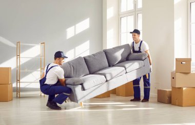 Oturma odasında ağır kanepe taşıyan iki genç servis çalışanı. Teslimat servisinden gelen erkek işçiler yeni eve mobilya yerleştiriyorlar. Taşınma günü ve taşınma konsepti.