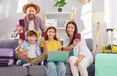 Pasaportları gösteren ve internetten bilet alan mutlu bir aile. Evdeki kanepede dizüstü bilgisayarla yaz tatili planlayan bavullarla oturuyor. Tatil gezisi konsepti.