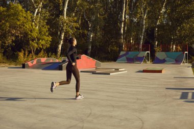 Spor sahasında koşan ve şehir parkında kaykay rampasında koşan genç ve güzel bir kadının arka görüntüsü. Spor, egzersiz, spor, sağlık, egzersiz konsepti