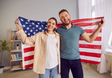 Amerikan gururu ve birliği. Arkalarında ikonik yıldız ve şeritlerle dikilen vatansever genç Amerikan çifti. Neşeli adam ve kadın evde, Amerikan bayrağı kameraya gülümsüyor..