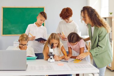 Öğretmen, ilkokulda biyoloji dersi sırasında kız öğrencinin mikroskoba nasıl baktığını gözlemliyor. Okul çocukları not defterlerine sınıf arkadaşlarının yazdırdığı bilgileri yazarlar.. 