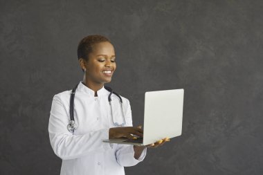 Mutlu gülümseyen doktorun dizüstü bilgisayardaki görüntüsü. Laboratuvar üniformalı kendine güvenen siyah kadın koyu gri arka planda, kablosuz internet bağlantısı olan modern bilgisayar kullanıyor.