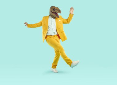 Dinozor maskesi takmış enerjik genç adam dans ediyor. Ürkütücü, tuhaf, sarı takım elbiseli bir adamın izole stüdyo arka planında eğlenirken tam boy fotoğrafı.