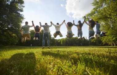 Yaz parkında eğlenirken havada el kaldıran bir grup arkadaş. Mavi gökyüzünün arka planında el ele tutuşan ve zıplayan insanların dikiz görüntüsü. Arka plan.