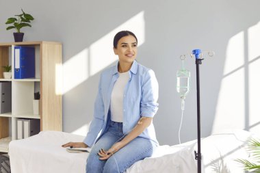 Tıbbi klinikteki kanepede oturup pencereye bakan genç ve mutlu bir kadın kanında serum ve vitamin enjeksiyonu terapisi görüyor. Sağlık hizmetleri kavramı.