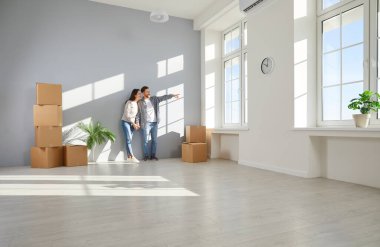 Yeni dairede karton kutularla duvarın önünde duran mutlu genç çift taşınma gününü kutlarken gülümsüyor ve pencereden manzaraya bakıyor. Taşınma, emlak, ipotek konsepti.