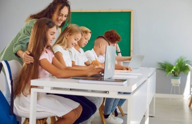 Okuldaki bir ders sırasında öğretmenler aktif olarak öğrencilere yardım ediyor, grup çalışmasını destekliyor. Çocuklar öğrenme teknolojisini sembolize eden bir dizüstü bilgisayarla uğraşıyorlar..