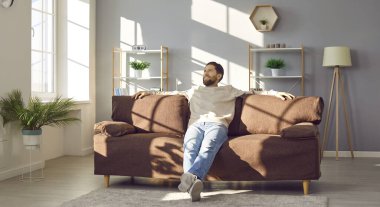 Evdeki yumuşak kanepede dinlenen yakışıklı genç adam. Modern, rahat, aydınlık oturma odasında kollarını açmış rahat koltukta oturan gülümseyen bir adam. Rahatlama zamanı, hayal kurma zamanı.
