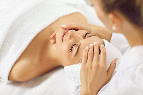 Beauty treatment skincare. Satisfied beautiful woman receiving facial treatment and professional massage while relaxing at spa. Close up of masseur smoothing face of woman lying on massage couch.
