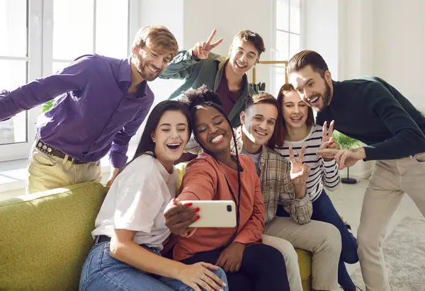 Happy young diverse group of friends students having fun enjoying party taking selfie with mobile phone sitting on sofa gathering at home together. Friendship and people lifestyle concept.