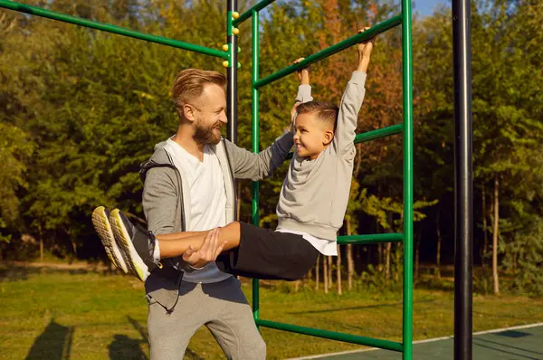 Mutlu gülümseyen bir baba çocuğuna spor için spor yaparken jimnastik barında egzersiz yaptırırken oğlu da açık havadaki spor malzemelerini sigortalarken yardım ediyor. Sağlıklı yaşam tarzı ve doğa konsepti eğitimi.