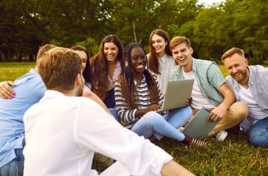 Bir grup mutlu, neşeli, neşeli, çeşitli uluslar arası, çoklu etnik çeşitlilikte üniversite öğrencisi arkadaşlar yeşil çimlerin üzerinde oturup birlikte çalışıyorlar, dizüstü bilgisayar kullanıyorlar, eğleniyorlar, komik şakalara gülüyorlar.