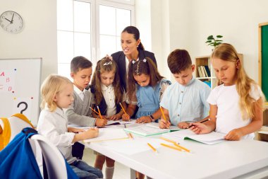 Bir grup ilkokul öğrencisi, genç bayan öğretmenle birlikte görevi tamamlıyor. Okul çocukları bir masada durur ve öğretmenlerini dinlerken not defterlerine yazarlar..