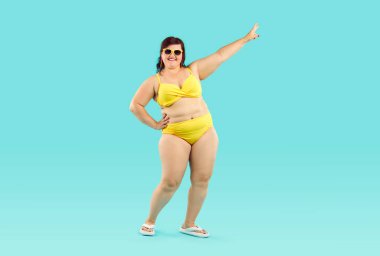 Sarı bikinili, güneş gözlüklü, yaz tatili gezisine çıkan ve mavi stüdyo arka planında eğlenen komik genç bir kadının vücut portresi. Tatil ve seyahat kavramı.