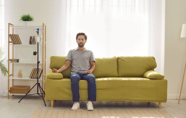 Evdeki ya da klinikteki kanepede oturan genç adam kanında serum ve vitamin tedavisi görürken. Günlük giysiler içinde üzgün bir erkek iğne terapisi görüyor..