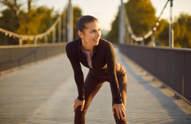 Şehir parkındaki spor salonunda güzel bir kadın. Modern spor kıyafetleriyle genç, zayıf, çekici bir kızın portresi köprüde dizlerinin üzerine çöküp başka tarafa bakıyor. Spor konsepti
