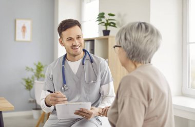 Dost canlısı doktor klinik randevusu sırasında yaşlı kadına bilgilendirici tıbbi danışmanlık yapıyor. Gülümseyen doktor olgun hastaya tedaviden bahsediyor ve muayene sonuçlarını açıklıyor.