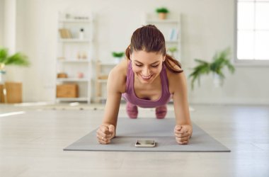 Mutlu, formda, sportif, gülümseyen genç bir kadın cep telefonunda fitness uygulaması kullanıyor ve evdeki spor salonunda egzersiz yaparken yoga minderi üzerinde dirsek dirseği egzersizi yapıyor.