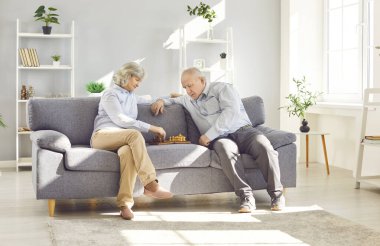 Gray-haired senior old couple playing chess together sitting on sofa in living room at home. Retired elderly man and woman playing board games enjoying retirement. Family leisure concept.
