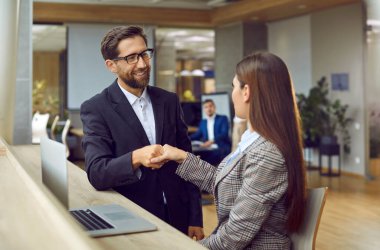 Two business people meet in the office, make a deal and exchange handshakes. Happy man and woman in suits sitting at a table with a laptop computer and shaking hands. Professional teamwork concept