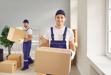 Portrait of smiling young man mover worker in uniform holding a cardboard box and looking cheerful at camera standing in empty new apartment. Move, relocation and moving service concept.