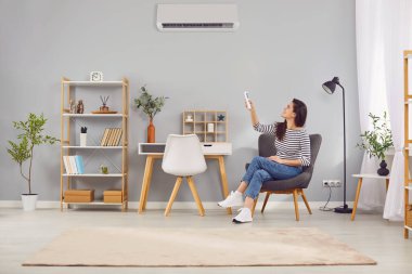 Young satisfied woman at home adjusts air conditioning in room using remote control. Woman sitting comfortably on chair in home office pointing remote control at air conditioner mounted on gray wall.