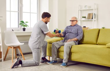 Senior man doing exercise using dumbbells with support from nurse.Young smiling caregiver physiotherapist helping elderly man in lifting dumbell at retirement. Recovery and nursering home concept.