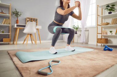 Young sporty woman doing stretching exercises with rubber band standing in living room at home. Happy athletic girl doing domestic workout. Sport, fitness and home training concept.