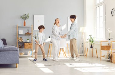 Happy smiling mother having fun and dancing with her two children boys in the living room at home. Young woman parent enjoying spending time with sons. Family leisure and mothers day concept.