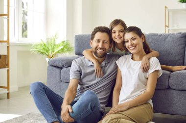 Cheerful smiling happy family sitting on the floor in the living room at home with their child girl and looking at camera. Cute daughter hugging her parents on sofa. Love and family leisure concept.